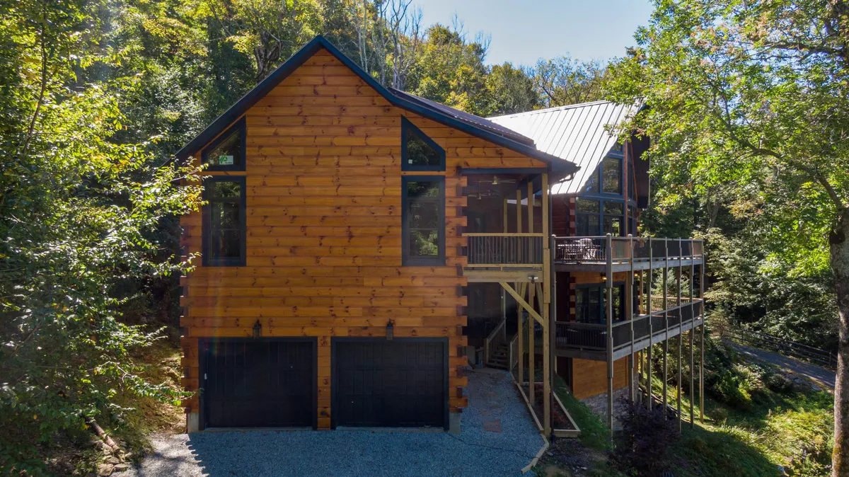 A welcoming exterior view of the luxury cabin, featuring a wooden facade with large windows, a spacious balcony, and trees surrounding the property. The scene captures sunset light and suggests possibilities for outdoor gatherings or relaxing on the balcony, with a backdrop of natural beauty.