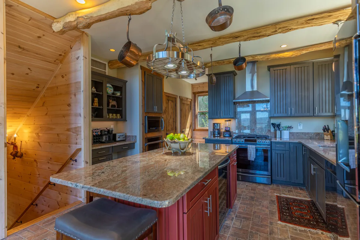 A bright, stylish kitchen with a prominent granite island, hanging light fixtures, wooden ceiling beams, and modern stainless steel appliances. The setup includes double ovens and ample prep space—ideal for both everyday cooking and celebratory feasts.