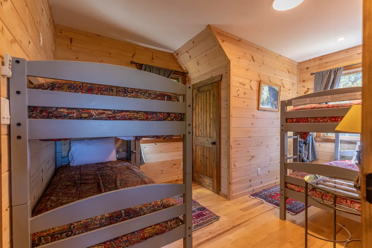 A warm and inviting bunk room with two twin-over-twin bunk beds, wood-paneled walls, a decorative rug, and a small table with a lamp. The setup is playful yet cozy, encouraging bonding, giggles, and sleepovers for children or friends.