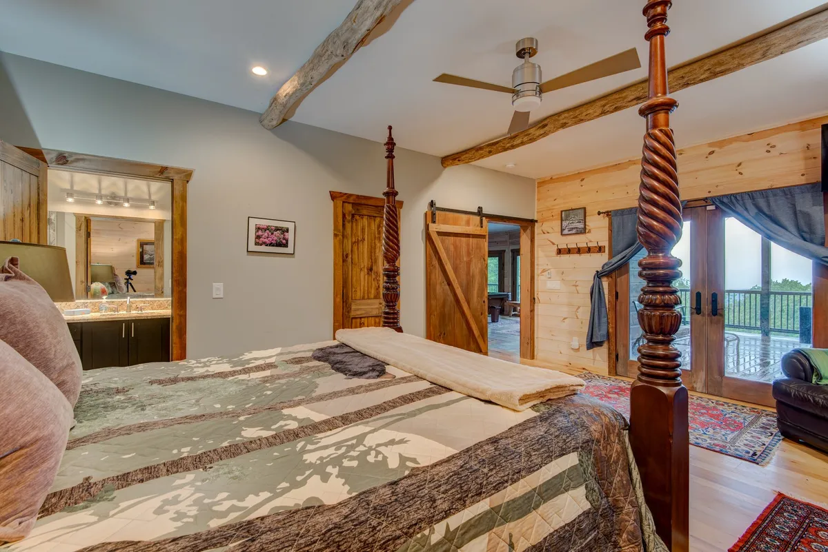 A serene bedroom featuring a four-poster bed with a decorative quilt, wooden beams overhead, sliding barn doors, and an outdoor view that hints at a Juliet balcony. Soft natural light brightens the space, creating an inviting atmosphere perfect for relaxation and private moments.