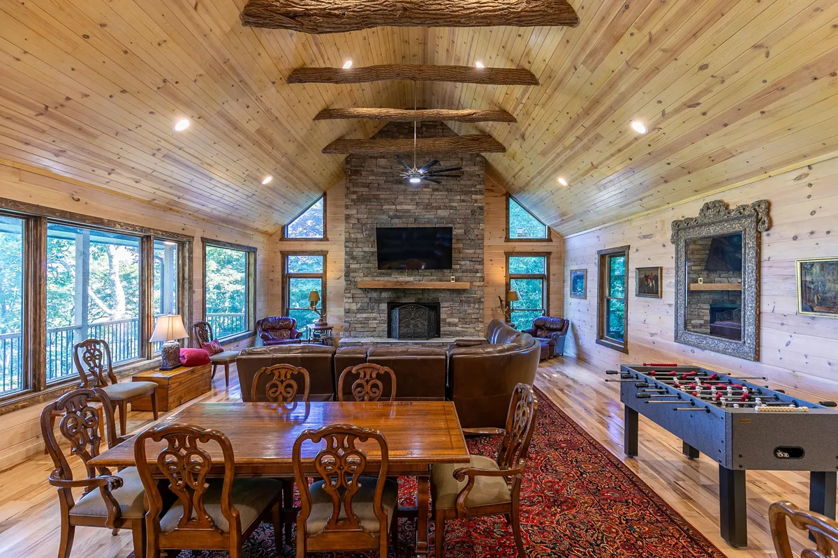A cozy, inviting interior scene featuring a large stone fireplace, dining table, foosball table, and leather sofas in a great room. Large windows reveal mountain views, while the space encourages family gatherings, board games, and relaxing evenings by the fire.