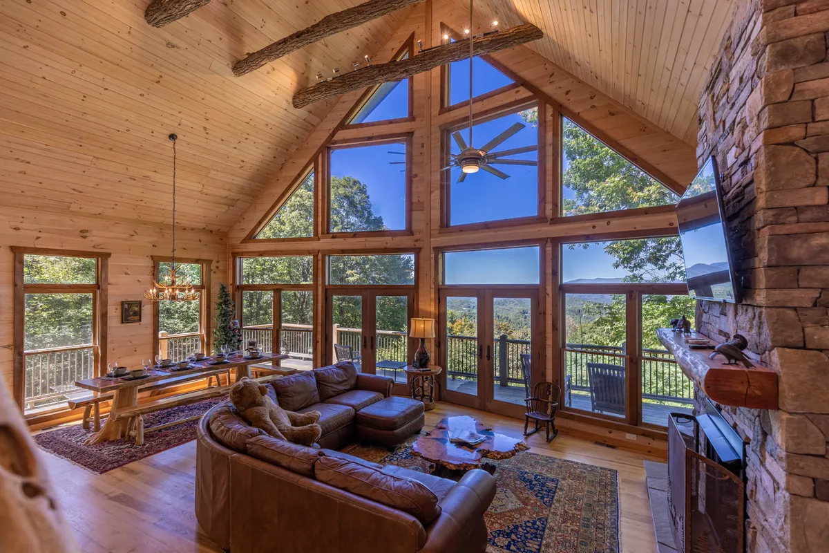 A welcoming interior scene showcasing large windows with awe-inspiring mountain views, rustic wooden beams, a plush leather sofa, a stone fireplace, and a dining table set for meals. The space exudes warmth and togetherness, with architectural features like vaulted ceilings and abundant natural light.