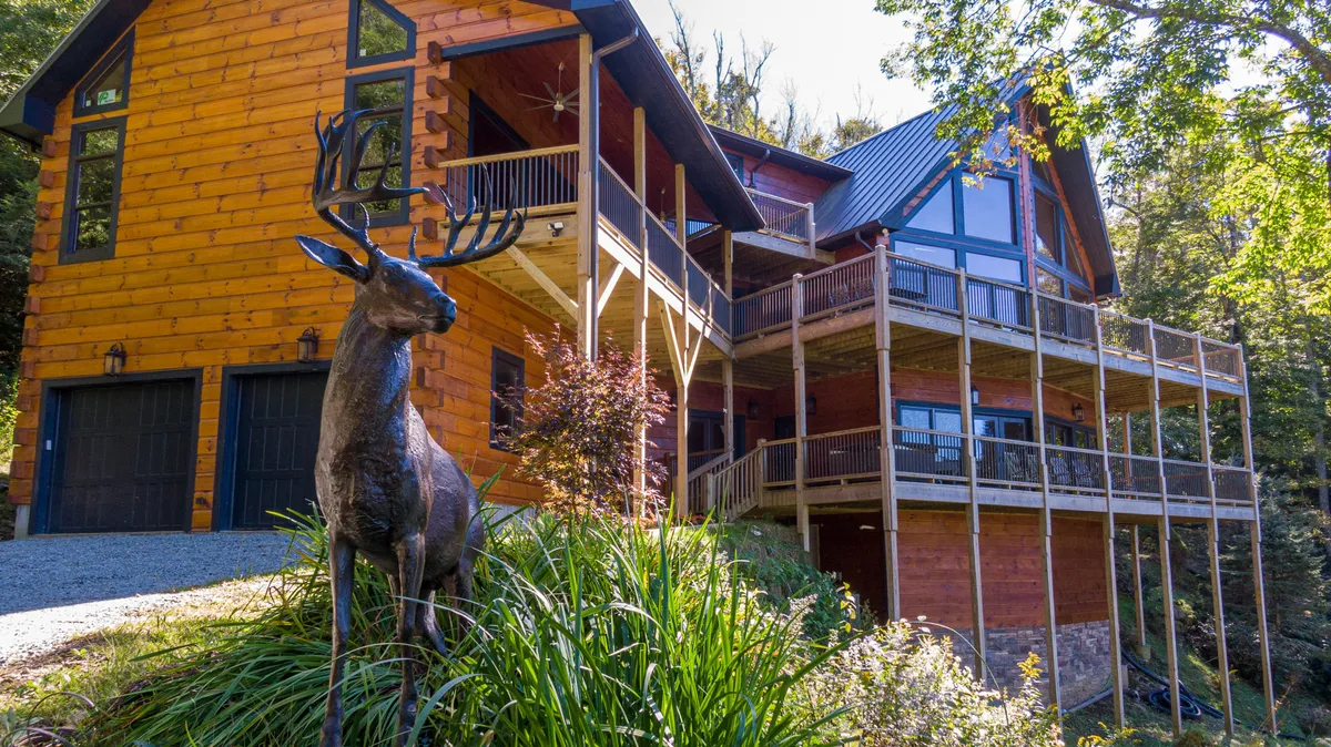 A welcoming exterior view of a large mountain house surrounded by lush greenery, with balconies and a prominent deer statue in the yard. The setting conveys a peaceful retreat nestled in nature, ideal for peaceful walks and outdoor exploration.