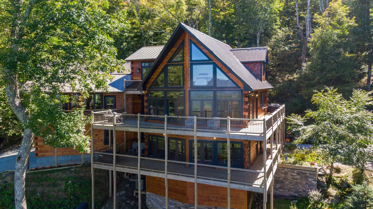 The image showcases the cabin’s striking wooden exterior with large glass windows set atop a sturdy stone foundation. An inviting deck wraps around the home, perfectly positioned for sunset views, outdoor gatherings, and relaxation surrounded by lush mountain scenery high on Appalachian Ski Mountain.
