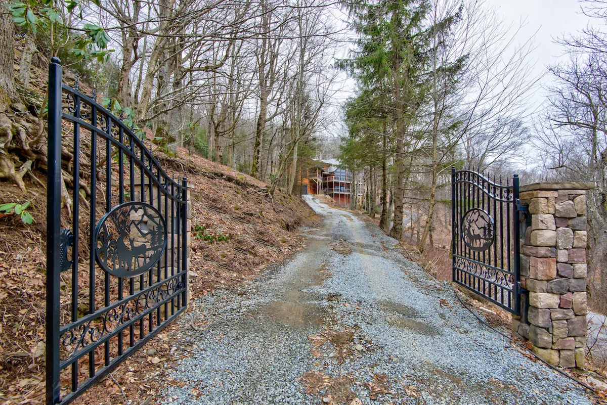 This exterior shot features ornate iron gates at the property entrance, a gravel driveway, and mature surrounding trees. It conveys a sense of arrival and sets the tone for an alpine retreat, while also alluding to the need for AWD/4WD vehicles during winter months as mentioned in the winter note.