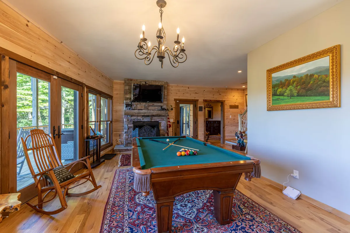 A warmly lit game room featuring a pool table at center stage, inviting seating including a rocking chair, a cozy fireplace, and a landscape painting on the wall. The ambiance is inviting and cozy, perfect for group games or relaxing by the fire.
