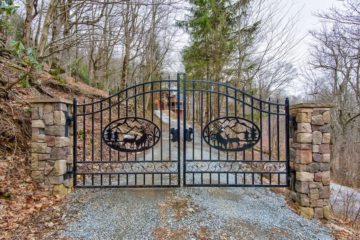An ornate metal gate with stone pillars marks the entry to the property, bordered by tall trees. The scene conveys an immediate sense of exclusivity and arrival into a secluded, high-end retreat.
