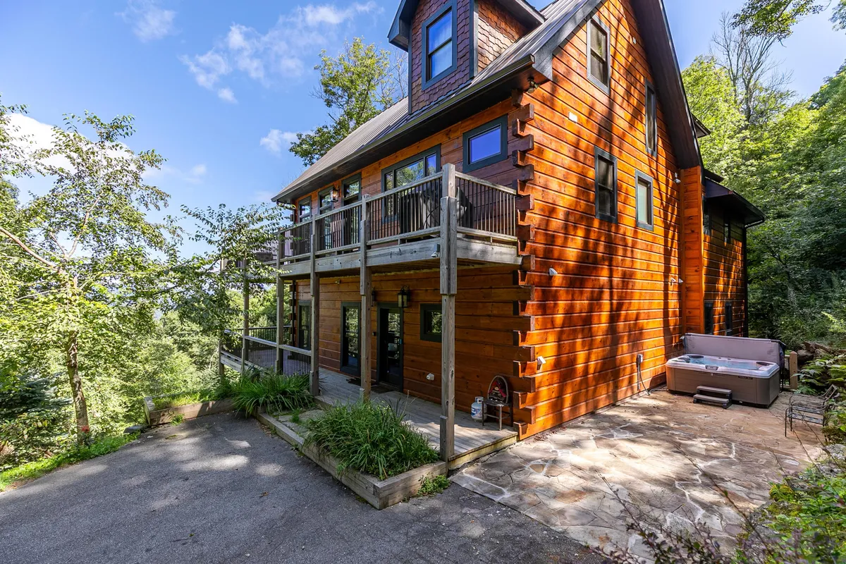 This exterior image captures the warm wooden facade of Rested Traveler, with a spacious balcony and the prominent 8-person Bullfrog Spas hot tub surrounded by tastefully landscaped outdoor areas. The scene suggests an inviting atmosphere for relaxing after a day of adventure, with natural elements and gathering spaces clearly visible.