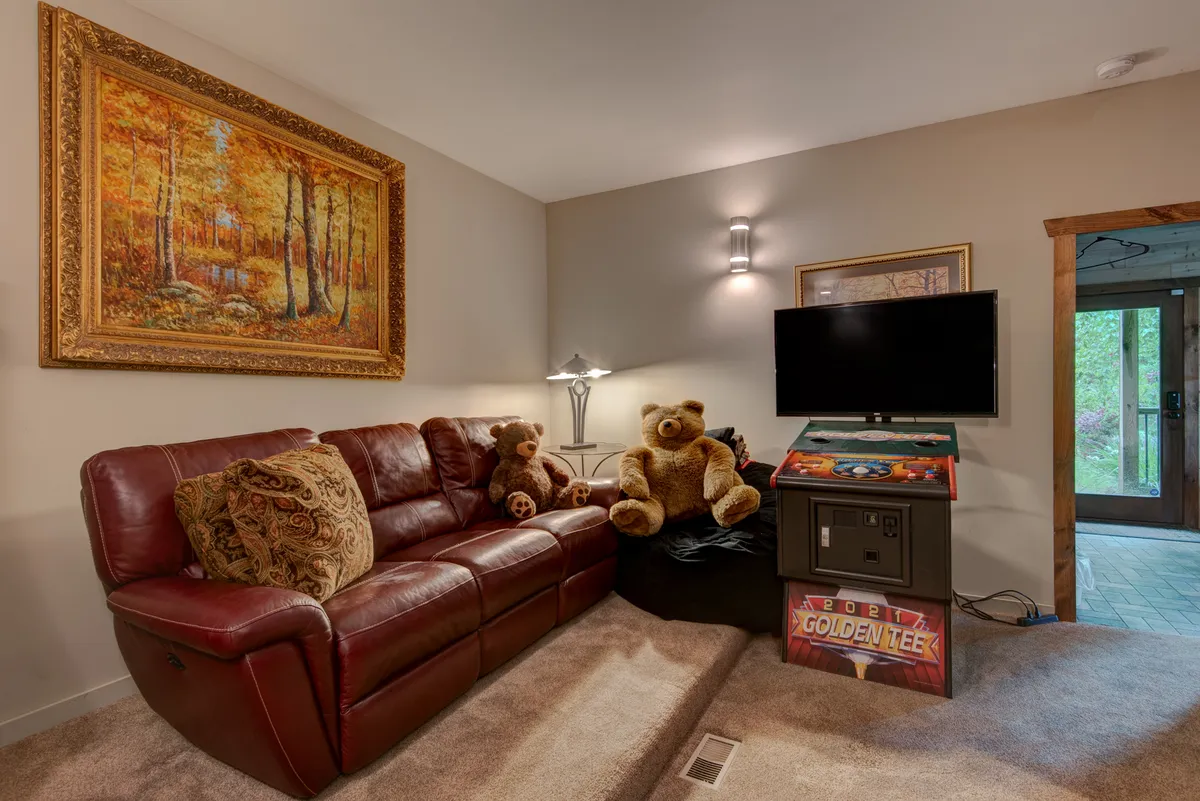 A playful entertainment space with a vibrant red leather sofa, a wall-mounted TV, an arcade game machine, and themed decor like teddy bears and a landscape painting. The atmosphere is both fun and comfortable, echoing the room’s use for games and movie nights.
