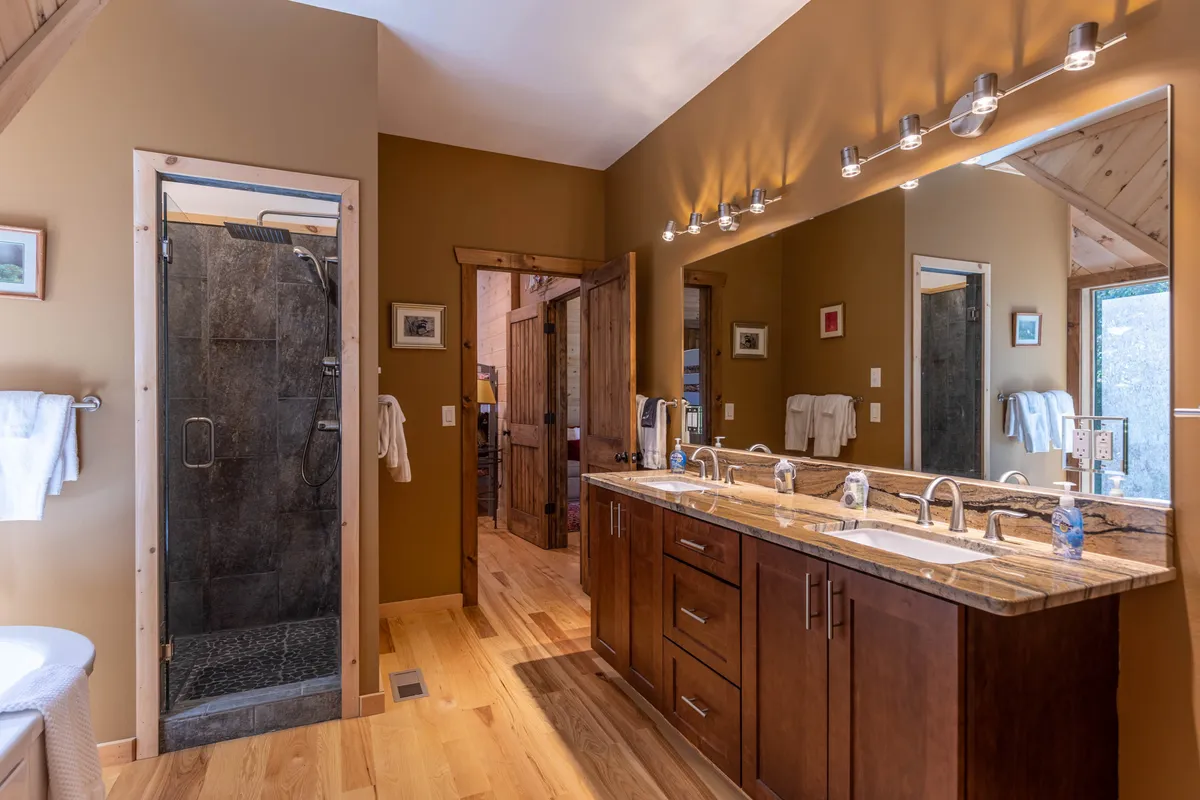 A modern shared bathroom showcasing a double granite vanity, a spacious walk-in shower with glass doors, and wooden accents for a spa-like ambiance. The mirrored wall expands the space and enhances light, creating a fresh and luxurious experience.