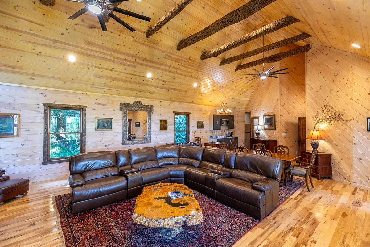 A spacious great room with a large leather sectional sofa, rustic tree-stump coffee table, decorative mirror, and prominent wooden ceiling beams. The layout encourages socializing, relaxation, or entertainment.